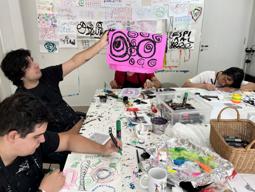 Criativos com deficiência intelectual do Estudio QUE sentados em uma grande mesa branca fazem colagens e pinturas em formas de espirais