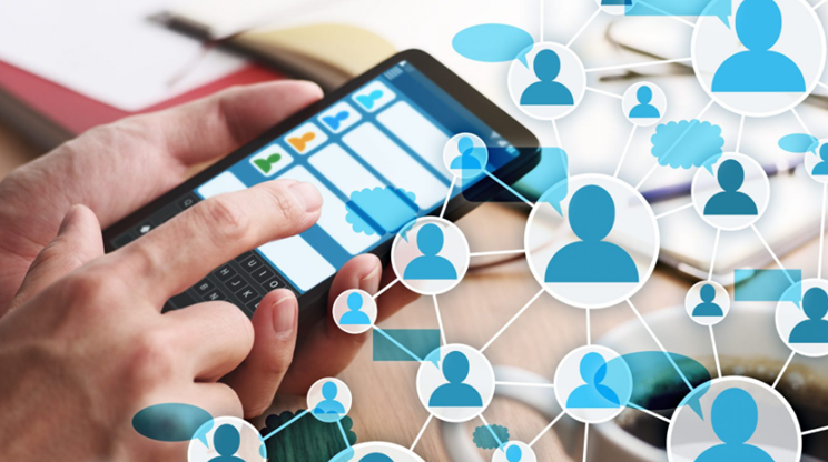 Foto das mãos de uma pessoa segurando um smartphone. Na tela há ícones de pessoas. Há figuras estilizadas sobrepostas na foto de perfis humanos conectadas por linhas brancas, simbolizando uma rede. O fundo contém objetos desfocados, como um caderno, uma caneta e uma xícara de café.